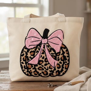 Op de afbeelding: Crèmekleurige draagtas met een pompoenontwerp. De pompoen heeft een luipaardprint met een roze strik en een zwarte steel. De tas heeft twee handvatten en staat op een houten oppervlak.