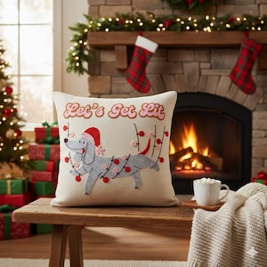 May include: A square, off-white decorative pillow with an embroidered dog wearing a Santa hat and Christmas lights. The text "Let's Get Lit" is embroidered above the dog. The pillow is on a wooden bench in front of a fireplace.
