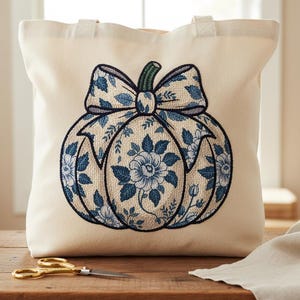 May include: Cream-colored tote bag featuring a blue floral pumpkin design with a bow. The pumpkin is embroidered with a detailed pattern of blue flowers and leaves. The bag has two handles and is sitting on a wooden surface.