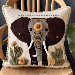 Peut inclure: Un coussin beige avec un motif d'éléphant brodé en marron, gris et jaune. L'éléphant a un motif floral sur la tête et est entouré de feuilles vertes et de fleurs jaunes. Le coussin a une bordure à franges et le texte "PATTERNSANDTHERAPY" est brodé en haut.