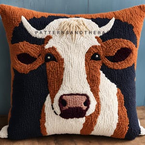 May include: A brown and white cow face is depicted on a plush pillow with a dark blue background. The pillow is made of yarn and has a textured, fuzzy appearance. The cow's eyes are black with white highlights, and its nose is pink. The text "PATTERNSANDTHERA" is visible in the upper center of the pillow.