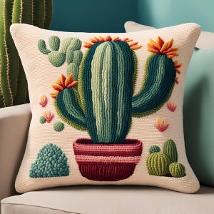 May include: A decorative square pillow with a cream-colored background features a detailed embroidered cactus design. The cactus is green with orange accents, set in a burgundy pot. Additional cacti and floral elements are also embroidered.
