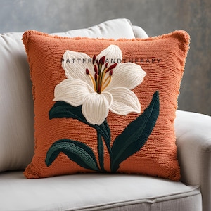 May include: A burnt orange throw pillow with a white embroidered lily flower and green leaves. The pillow has a fringed edge.