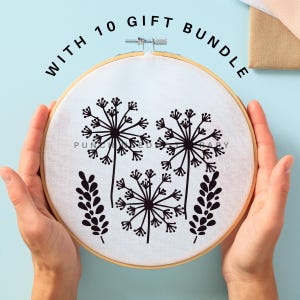 May include: A white embroidery hoop with black embroidery of stylized dandelions and leaves. The hoop is held in a person's hands. The text "WITH 10 GIFT BUNDLE" is written in black around the hoop.