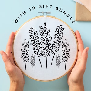 May include: A close-up of a white embroidery hoop with a black floral design. The hoop is being held by a person's hands. The text "WITH 10 GIFT BUNDLE" is written around the hoop.