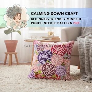 Könnte beinhalten: Ein dekoratives Kissen mit einem Blumendesign aus roten, lila und rosa Rosen. Das Kissen liegt auf einem weißen Teppich vor einem beigen Sessel. Der Text "CALMING DOWN CRAFT" und "BEGINNER-FRIENDLY MINDFUL PUNCH NEEDLE PATTERN PDF" ist sichtbar.