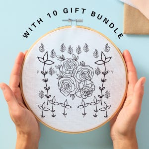 May include: A white embroidery hoop with a black line drawing of a floral design. The hoop is held in the hands of a person. The text "WITH 10 GIFT BUNDLE" is written in black letters around the hoop.