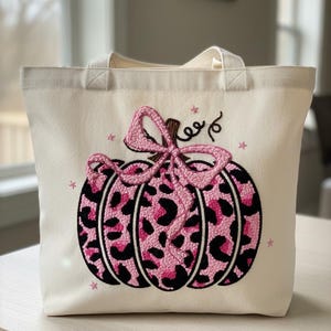 Könnte beinhalten: Cremefarbene Tragetasche mit einem gestickten Kürbis-Design mit rosa Leopardenmuster. Der Kürbis hat eine rosa Schleife und ist von kleinen rosa Sternen umgeben. Die Tasche hat zwei Griffe.