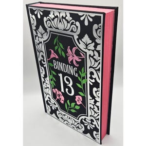 May include: A decorative book with a black cover featuring silver floral patterns and the text "BINDING 13". The book's spine and edges are pink. The cover also has pink and green floral accents.