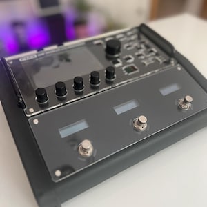 Peut inclure: Une pédale d'effets multiples noire avec un grand écran LCD, quatre boutons et deux commutateurs au pied. La pédale a une garniture argentée et un couvercle en plastique transparent.