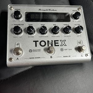 Op de afbeelding: Een zilveren ToneX gitaareffectpedaal met een zwart-wit logo. Het pedaal heeft een digitaal display, knoppen voor gain, bas, mid, treble, volume, reverb, compressor, noise gate en presence, en drie voetschakelaars met de labels A, B en C. De voetschakelaars zijn gelabeld "Bank Down", "Bank Up" en "VIR™ Tech".