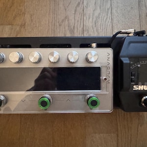 Puede incluir: Un dispositivo de audio plateado y transparente con múltiples perillas y una pantalla, junto a un dispositivo negro con una pantalla y la palabra "SHURE". El dispositivo tiene la palabra "ANAGRAM" impresa.