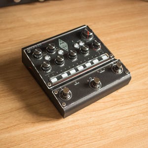 Può includere: Un pedale per effetti per chitarra nero con numerosi pulsanti e interruttori. Il pedale ha una superficie strutturata e dettagli argentati. La parola "PROFILER" è stampata in alto a sinistra. Il pedale è su una superficie di legno.