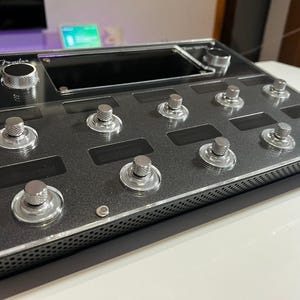 Peut inclure: Une pédale d'effets de guitare Fender noire avec un écran numérique et huit commutateurs au pied. La pédale a un bouton argenté et un design noir et argent.