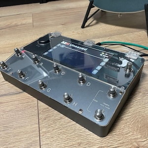 May include: A black and silver multi-effect pedal with a clear plastic cover. The pedal has eight footswitches labelled A through H, a knob, and a screen that reads "32H Unsaved".