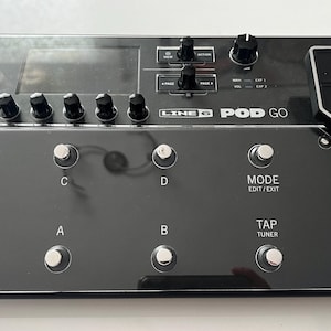 Könnte beinhalten: Ein schwarzes Line 6 POD Go Gitarreneffektpedal mit einer transparenten Plastikabdeckung. Das Pedal verfügt über eine Vielzahl von Knöpfen und Reglern, darunter ein Bildschirm, ein Tap-Tuner und ein Modus-Knopf.