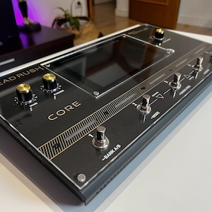 Può includere: Un processore di effetti per chitarra Headrush Core nero con un piccolo schermo, due manopole e quattro pedali. I pedali sono etichettati "Mode", "Harmonic", "Pre-Delay" e "Bank A/B".