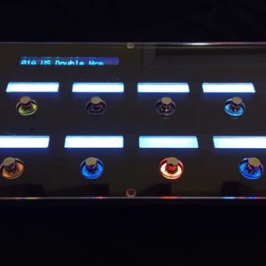 Puede incluir: Un dispositivo electrónico negro con múltiples botones y pantallas iluminadas. El dispositivo tiene una parte superior de acrílico transparente. Los botones están iluminados con varios colores, incluyendo azul, amarillo y rojo. La pantalla muestra texto.