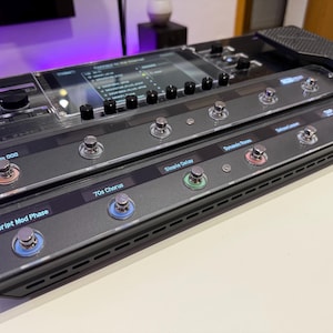 Pode incluir: Um processador de efeitos preto e cinza com vários pedais e um ecrã. O dispositivo tem rótulos como "Scream OD", "70s Chorus" e "Simple Delay". O dispositivo foi concebido para uso com instrumentos musicais.