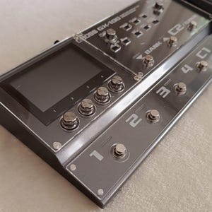 Pode incluir: Um pedal de efeitos múltiplos preto com um visor digital e cinco pedais comutadores etiquetados de 1 a 5. O pedal tem uma cobertura de plástico transparente e o texto "BOSS GT-1000 CORE" é visível na parte superior.
