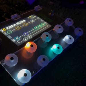 Può includere: Primo piano di un dispositivo elettronico nero con la scritta "1C KORA KORA" sullo schermo. Presenta diverse manopole traslucide illuminate in arancione, blu e bianco. Lo sfondo è scuro.