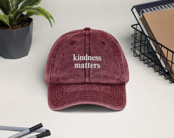 May include: A maroon vintage washed baseball cap with white embroidered text that reads "kindness matters".