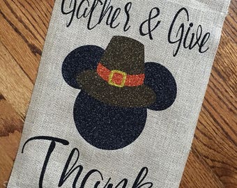 May include: A burlap garden flag with the words "Gather & Give Thanks" in black script. A black silhouette of a character's head is topped with a brown pilgrim hat with a red and gold buckle. The flag is rectangular.