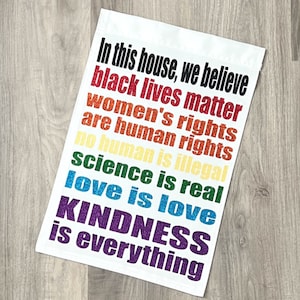 May include: White rectangular flag with text in various colors. The text reads "In this house, we believe" followed by statements about human rights, science, and love. The flag also includes the text "Dainty Diva Bows".