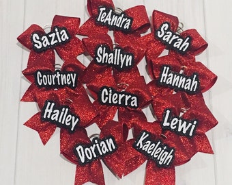 May include: A collection of red glitter hair bows, each with a name in white lettering on a black background. The bows are arranged in a cluster, with the text "Dainty Diva Bows" at the bottom.