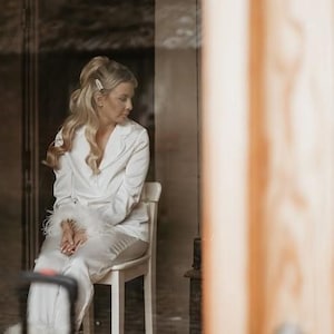 May include: A woman wearing a white satin pajama set with feather trim. The set includes a long-sleeved shirt and pants. The woman is sitting on a white stool.