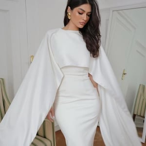 Peut inclure: Une robe blanche avec une longue cape. La robe a un corsage ajusté et une jupe ample. La cape est attachée aux épaules et tombe jusqu'au sol.