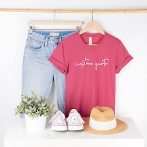 K&ouml;nnte beinhalten: Ein rotes T-Shirt mit dem Text "custom quote" in wei&szlig;en Buchstaben. Das Shirt wird mit einer blauen Jeans, wei&szlig;en Turnschuhen und einem Strohhut kombiniert.