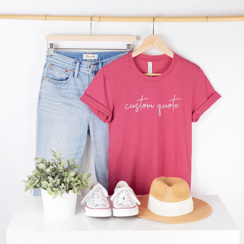 K&ouml;nnte beinhalten: Ein rotes T-Shirt mit dem Text "custom quote" in wei&szlig;en Buchstaben. Das Shirt wird mit einer blauen Jeans, wei&szlig;en Turnschuhen und einem Strohhut kombiniert.