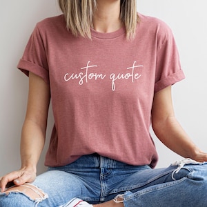 K&ouml;nnte beinhalten: Eine Frau tr&auml;gt ein heidefarbenes T-Shirt mit dem Text "custom quote" in wei&szlig;en Buchstaben. Sie tr&auml;gt eine blaue Jeans und sitzt mit gekreuzten Beinen.
