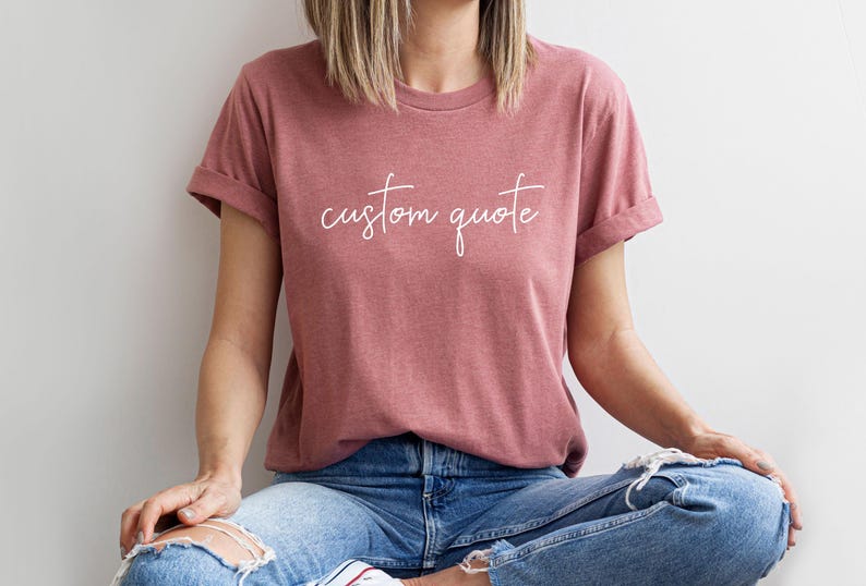 K&ouml;nnte beinhalten: Eine Frau tr&auml;gt ein heidefarbenes T-Shirt mit dem Text "custom quote" in wei&szlig;en Buchstaben. Sie tr&auml;gt eine blaue Jeans und sitzt mit gekreuzten Beinen.