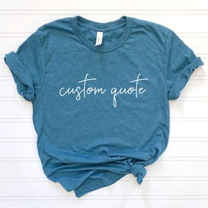 K&ouml;nnte beinhalten: Ein blaues T-Shirt mit dem Text "custom quote" in wei&szlig;en Buchstaben.