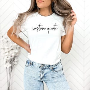 K&ouml;nnte beinhalten: Ein wei&szlig;es T-Shirt mit dem Text "custom quote" in schwarzer Kursivschrift.