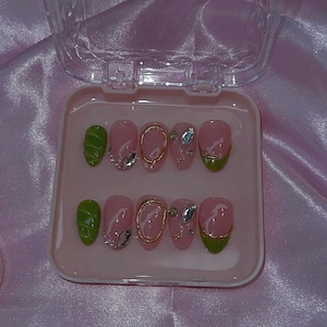 May include: A set of pink press-on nails with gold rings, rhinestones, and green tips, displayed in a clear plastic case. The nails are arranged on a pink background.