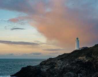 Könnte beinhalten: Ein weißer Leuchtturm steht hoch auf einer felsigen Klippe und überblickt ein blaues Meer. Der Himmel ist ein wunderschöner Mix aus Rosa und Blau mit zarten Wolken.