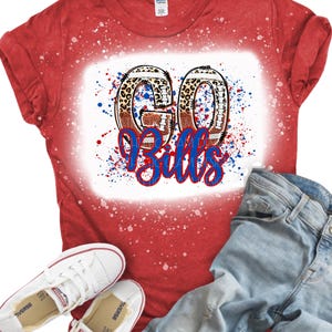 May include: A red short-sleeved t-shirt with a bleached effect, featuring a graphic of two footballs shaped like the letters G and O, with the text "Bills" in blue script below, paired with light blue denim jeans and white canvas sneakers.