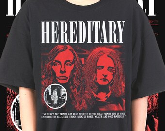 May include: Black t-shirt with the word "HEREDITARY" in white block letters. The shirt features a red graphic of two faces and a circular image. Below the graphic is text that reads "WE REJECT THE TRINITY..."