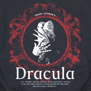 May include: Black t-shirt featuring a red framed design with the text "Bram Stoker's" at the top. The design includes a white illustration of a figure with hands covering the face, and the word "Dracula" in white.