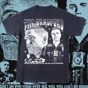 May include: Dark navy blue t-shirt featuring a graphic of two men's portraits and the text "TIM BRADFORD" in white. The shirt also includes the quote, "You don't let everyone ever tell you you can't do something."