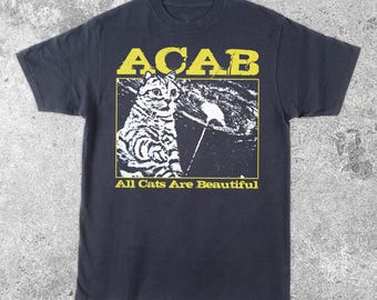 May include: Dark navy blue t-shirt with a yellow and white graphic. The graphic features the text "ACAB" above a black and white illustration of a cat. Below the cat illustration is the text "All Cats Are Beautiful".