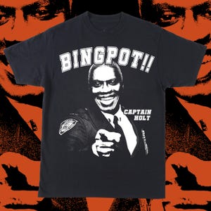 Puede incluir: Camiseta negra con una imagen en blanco y negro de un hombre sonriente con uniforme de policía señalando. El texto "BINGPOT!!" está encima de la imagen, con "CAPTAIN HOLT" debajo.