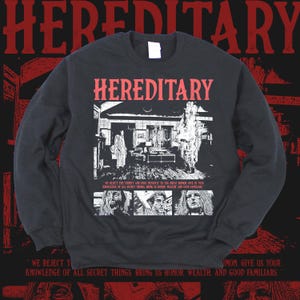 K&ouml;nnte beinhalten: Schwarzes Sweatshirt mit rotem Schriftzug "HEREDITARY". Die Vorderseite zeigt eine Schwarz-Wei&szlig;-Illustration eines Hausinneren und Gesichter. Der Text unten lautet "WE REJECT THE TRINITY..." und "SIMON GIVE US YOUR..."