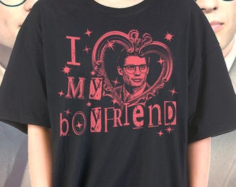 May include: Black t-shirt with a red graphic that says "I MY boyfriend". The graphic includes a portrait of a man wearing glasses inside a heart-shaped frame, surrounded by stars. The shirt is a casual style.
