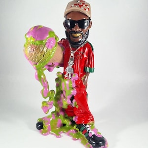 Puede incluir: Una figurita roja y verde de una persona que lleva un sombrero negro, gafas de sol y una cadena de oro. La persona sostiene un cono de helado rosa y verde. El helado se derrite y gotea por las piernas de la persona.