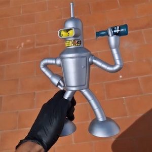 May include: A silver robot toy holding a blue bottle with the text "Bite My Shiny Metal A**" printed on it. The robot has yellow eyes and a silver body.