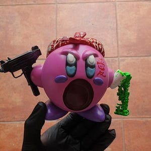 Puede incluir: Un personaje rosa con una boca grande, que lleva un pañuelo rojo con la palabra "Yakuza" escrita en él, sosteniendo una pistola negra en una mano y una cadena verde en la otra.